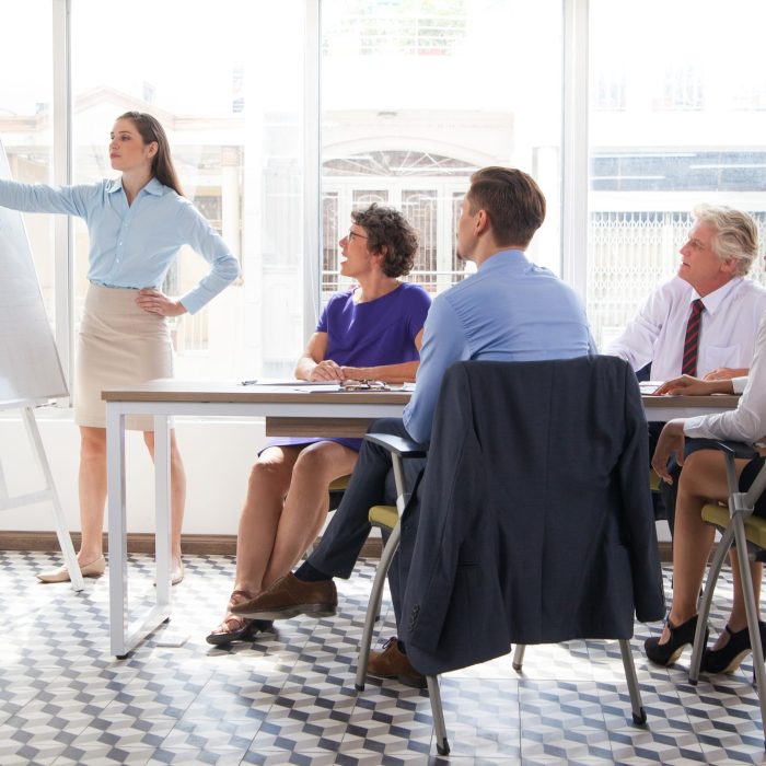 Confident female business coach pointing at whiteboard and explaining graph at meeting, group of four people listening to her. They sitting in conference room. Business training or education concept
